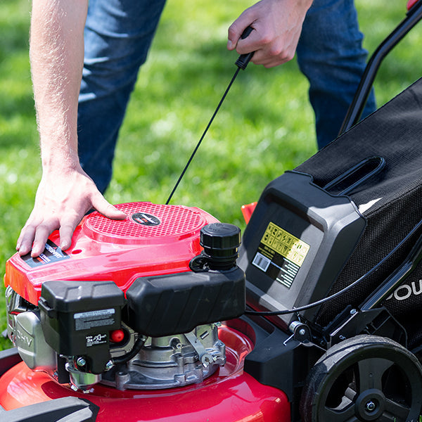 A close-up outdoor shot of a person using a red and black lawnmower, with one hand pulling the engine start cord and the other resting on the mower deck. The background shows a blurred green grass lawn.